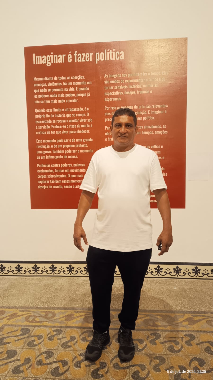 Imagem de um homem negro em uma galeria de arte em frente a uma placa escrito 'Imaginar é fazer política' com camisa branca e calças pretas